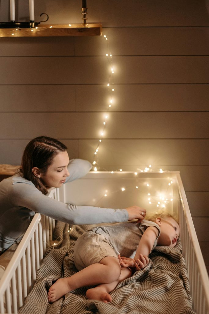 pexels-photo-7938230-7938230-1 A caring mother tends to her baby in a cozy crib illuminated by soft fairy lights.